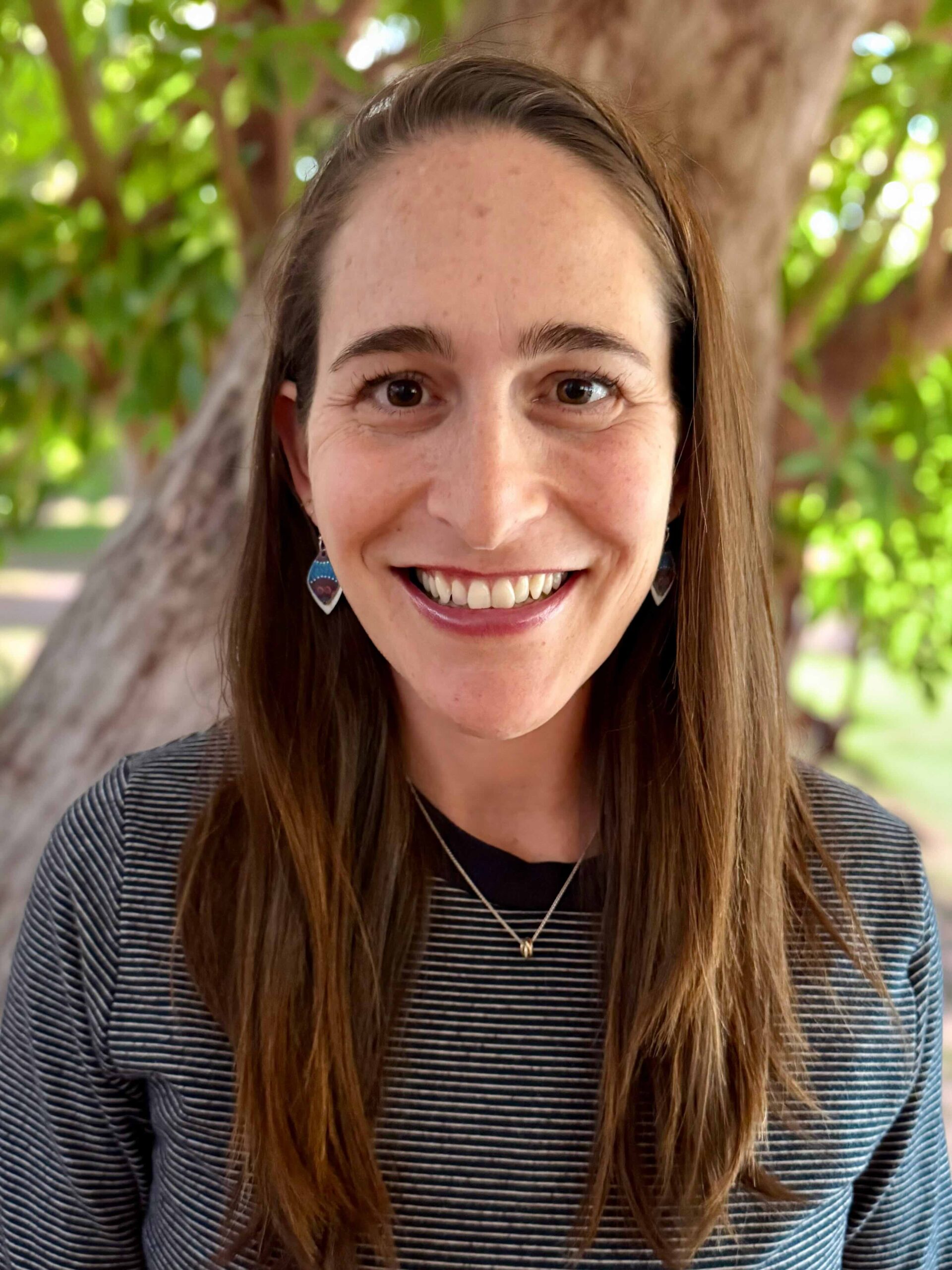 Close-up portrait of a smiling woman with long brown hair, wearing a striped shirt and heart-shaped earrings, outdoors by a tree.